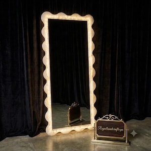 May include: A tall, rectangular floor mirror featuring a wavy, scalloped frame with a warm, illuminated glow. Beside it sits a small, rectangular metal sign with a dark wood center, displaying the gold script text Royalhandicraftexpo.