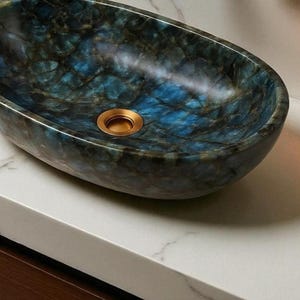 Pode incluir: Um lavatório oval com um padrão de pedra em azul profundo e cinza carvão com acabamento polido. A bacia apresenta um interior liso e arredondado com uma abertura de drenagem circular central num tom dourado metálico.