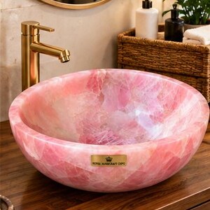 Pode incluir: Uma cuba redonda feita de pedra de quartzo rosa polido, com interior e exterior lisos. Na parte frontal, h&aacute; uma pequena placa met&aacute;lica retangular dourada com o texto ROYAL NAHHCRAFT DEPO e um &iacute;cone de coroa.