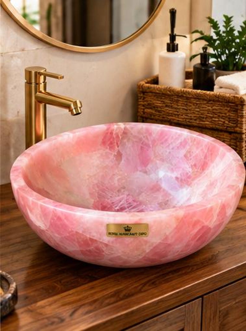 Pode incluir: Uma cuba redonda feita de pedra de quartzo rosa polido, com interior e exterior lisos. Na parte frontal, h&aacute; uma pequena placa met&aacute;lica retangular dourada com o texto ROYAL NAHHCRAFT DEPO e um &iacute;cone de coroa.