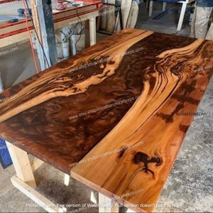 Op de afbeelding: Een groot, rechthoekig houten tafelblad met een natuurlijke houten afwerking. Het tafelblad heeft een donkerbruine epoxyharsinleg die door het midden van de tafel loopt. Het tafelblad wordt ondersteund door twee houten poten.