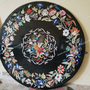 May include: A black marble table top with a floral design made of inlaid stones. The design features a variety of colors, including red, blue, green, and yellow. The table top is round and has a diameter of approximately 30 inches.
