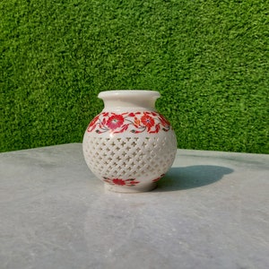 May include: White marble vase with a red floral design and intricate cut-out details. The vase has a round shape and a smooth, polished finish.