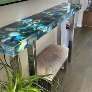 May include: A modern console table with a unique agate stone top. The table has a blue, green, and white agate pattern with a polished finish. The table is supported by two silver metal legs.