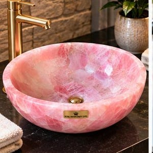 Pode incluir: Uma cuba redonda de quartzo rosa polido com uma textura cristalina lisa e transl&uacute;cida e um ralo dourado. Uma pequena placa retangular dourada na parte frontal exibe o texto Royal Handcraft Expo.