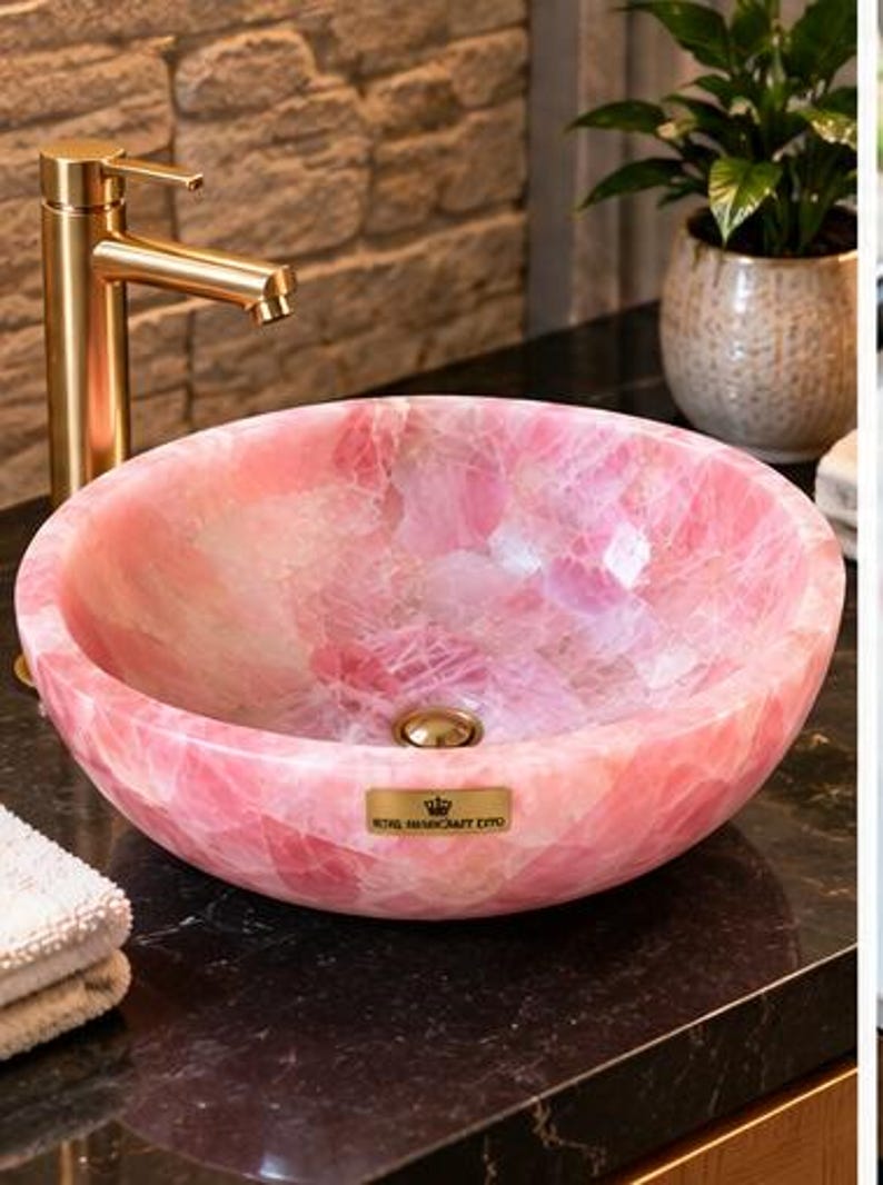 Pode incluir: Uma cuba redonda de quartzo rosa polido com uma textura cristalina lisa e transl&uacute;cida e um ralo dourado. Uma pequena placa retangular dourada na parte frontal exibe o texto Royal Handcraft Expo.