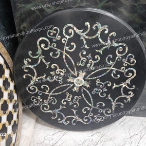 Puede incluir: Una tapa de mesa redonda negra con un diseño decorativo hecho de nácar incrustado. El diseño presenta un patrón floral con espirales y hojas. La tapa de la mesa tiene un diámetro de aproximadamente 61 cm.