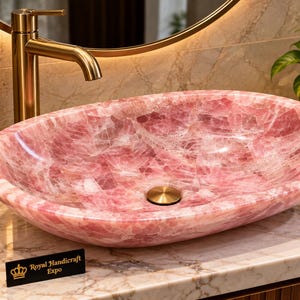 Pode incluir: Uma pia de banheiro oval esculpida em pedra de quartzo rosa translúcida com acabamento polido. A bacia possui um ralo central em tom dourado e repousa sobre uma bancada de mármore ao lado de uma pequena placa preta com os dizeres Royal Handicraft Expo.