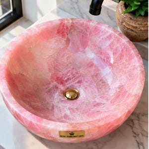 Pode incluir: Uma cuba redonda feita de pedra de quartzo rosa transl&uacute;cido com acabamento polido. A bacia apresenta um ralo em tom dourado e uma pequena placa retangular dourada na frente com o texto Royal Luminosity Expo.
