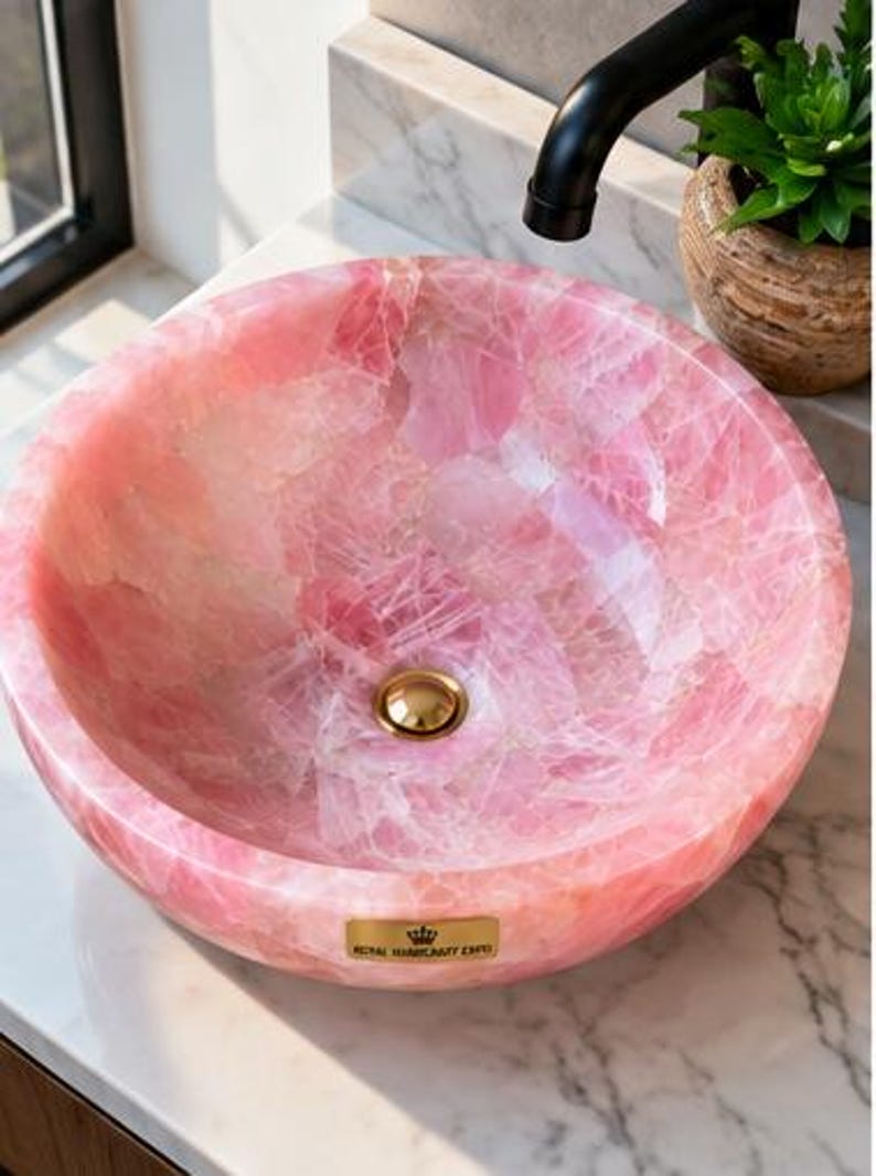Pode incluir: Uma cuba redonda feita de pedra de quartzo rosa transl&uacute;cido com acabamento polido. A bacia apresenta um ralo em tom dourado e uma pequena placa retangular dourada na frente com o texto Royal Luminosity Expo.
