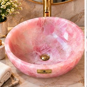 Pode incluir: Uma cuba de apoio redonda feita de pedra de quartzo rosa transl&uacute;cido com acabamento polido. A bacia apresenta um ralo em tom dourado e uma pequena placa retangular dourada na frente com o texto Royal Handicraft Corp.