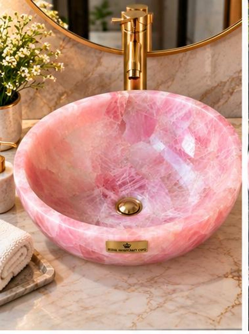 Pode incluir: Uma cuba de apoio redonda feita de pedra de quartzo rosa transl&uacute;cido com acabamento polido. A bacia apresenta um ralo em tom dourado e uma pequena placa retangular dourada na frente com o texto Royal Handicraft Corp.
