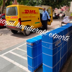 May include: Two large blue boxes with white tape and labels are being loaded into a yellow DHL delivery van. The boxes are stacked on top of each other. The van has the DHL logo and the words "Excellence Simple Deliveries" on the side.
