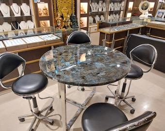 May include: A round table with a polished blue and gray labradorite stone top and a silver metal base, accompanied by four black swivel stools with silver metal frames and wheels, set in a retail environment with jewelry displays.