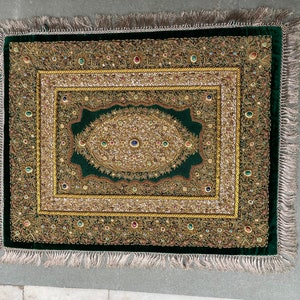 May include: A green velvet rug with a gold and jewel-toned embroidered design. The rug has a fringe border.