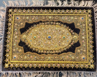 Puede incluir: Un textil rectangular de terciopelo negro con intrincados bordados de hilo metálico dorado en un patrón floral. El diseño incluye un motivo ovalado central adornado con pequeñas cuentas de colores y una piedra azul grande, con borde de flecos.