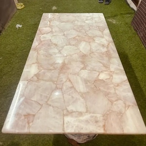 May include: Rectangular table with a polished, light pink and white stone mosaic top. The mosaic pattern is made up of irregular, interlocking pieces. The table is set on a neutral-colored base, and the surface reflects light.