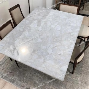 May include: A white marble dining table with a unique, cloudy pattern. The table is surrounded by four chairs with dark wood frames and light-colored upholstery. The table is set on a gray rug.