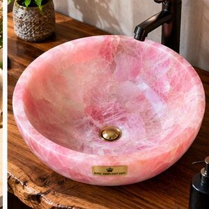 Pode incluir: Uma cuba de apoio redonda feita de pedra de quartzo rosa transl&uacute;cido com acabamento polido. A bacia apresenta um ralo em tom dourado e uma pequena placa retangular de lat&atilde;o com o texto ROYAL HANDICRAFT EXPO na borda frontal.