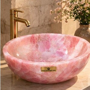 Pode incluir: Uma cuba redonda feita de pedra de quartzo rosa transl&uacute;cida com acabamento polido. Uma torneira de metal dourado fica atr&aacute;s da bacia. Uma pequena placa retangular dourada na frente apresenta um &iacute;cone de coroa e o texto ROYAL MAGICRAFT EXPO.