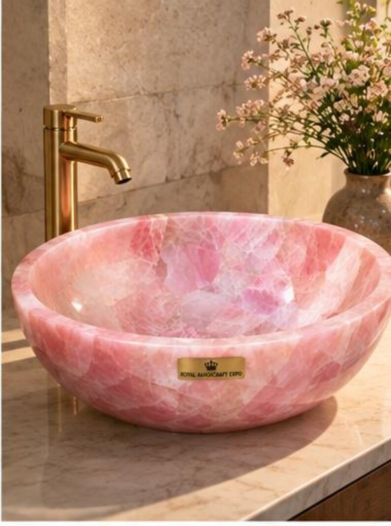Pode incluir: Uma cuba redonda feita de pedra de quartzo rosa transl&uacute;cida com acabamento polido. Uma torneira de metal dourado fica atr&aacute;s da bacia. Uma pequena placa retangular dourada na frente apresenta um &iacute;cone de coroa e o texto ROYAL MAGICRAFT EXPO.
