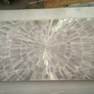 May include: A white mother of pearl inlay table top with a circular pattern. The table top is made of small pieces of mother of pearl that are arranged in a circular pattern. The table top is surrounded by a white border.