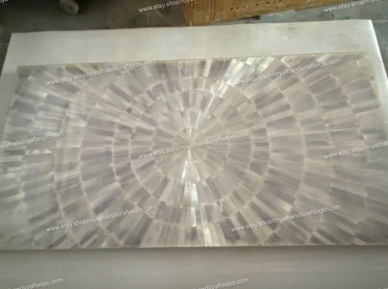 May include: A white mother of pearl inlay table top with a circular pattern. The table top is made of small pieces of mother of pearl that are arranged in a circular pattern. The table top is surrounded by a white border.