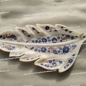 Puede incluir: Un plato de servir de mármol blanco con forma de hoja con intrincados diseños florales azules y dorados. El plato está decorado con piedras incrustadas y tiene un borde elevado.