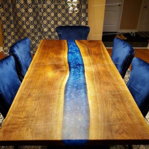 May include: A large wooden dining table with a blue epoxy river running down the center. The table is surrounded by blue velvet chairs.