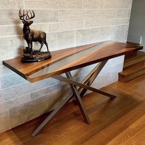 Puede incluir: Una mesa consola moderna con una tapa de madera de nogal y un diseño de río de epoxi plateado. La mesa tiene una base de metal en forma de X única.