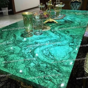 Puede incluir: Una gran mesa de malachite verde con detalles dorados. La superficie de la mesa está altamente pulida y tiene un patrón de remolino. La superficie de la mesa está rodeada de detalles dorados y un candelabro dorado.
