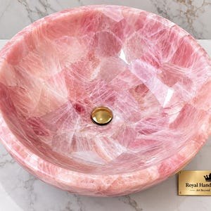 Pode incluir: Uma cuba redonda feita de quartzo rosa transl&uacute;cido com acabamento polido e um ralo central em tom dourado. Uma pequena placa retangular dourada est&aacute; presente com o texto Royal Handicraft Expo Art Beyond Boundaries.