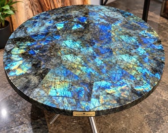 May include: A round side table featuring a polished labradorite stone top with vibrant blue, teal, and gold iridescent flashes set in a mosaic pattern. The table has a small gold-toned plaque labeled Royal Handcraft Expo and three metal legs.