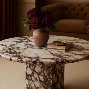 May include: A round coffee table with a marble top featuring a white and burgundy pattern. The table has a cylindrical base. A vase with burgundy flowers, a small gold bowl, and a book are on the table.