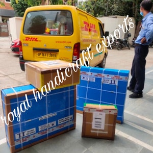 May include: A yellow DHL delivery truck with a blue and white box labeled "royal handicraft exports" on the back. The truck is parked in front of a building with a man standing next to it. There are several boxes on the ground in front of the truck.