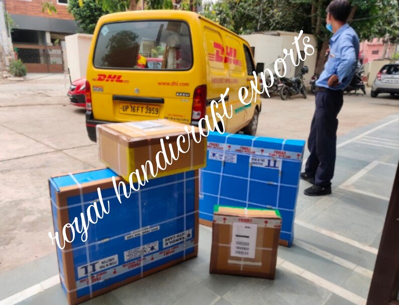 May include: A yellow DHL delivery truck with a blue and white box labeled "royal handicraft exports" on the back. The truck is parked in front of a building with a man standing next to it. There are several boxes on the ground in front of the truck.