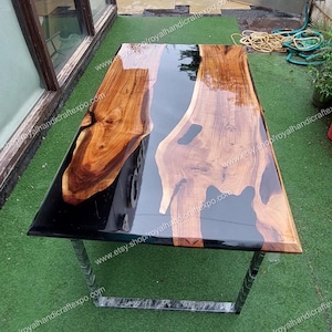 Op de afbeelding: Een houten tafelblad met een zwarte epoxyharsrivier die er doorheen loopt. De tafel heeft een natuurlijke houten afwerking en wordt ondersteund door een zwart metalen frame. De tafel is ongeveer 152 cm lang en 76 cm breed.  www.etsy.shop/royalhandicraftexpo.com