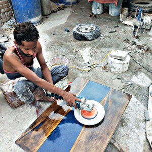 May include: A person uses a power sander to polish a wooden table top with a blue epoxy resin inlay. The table top is made of dark wood with a natural grain pattern.