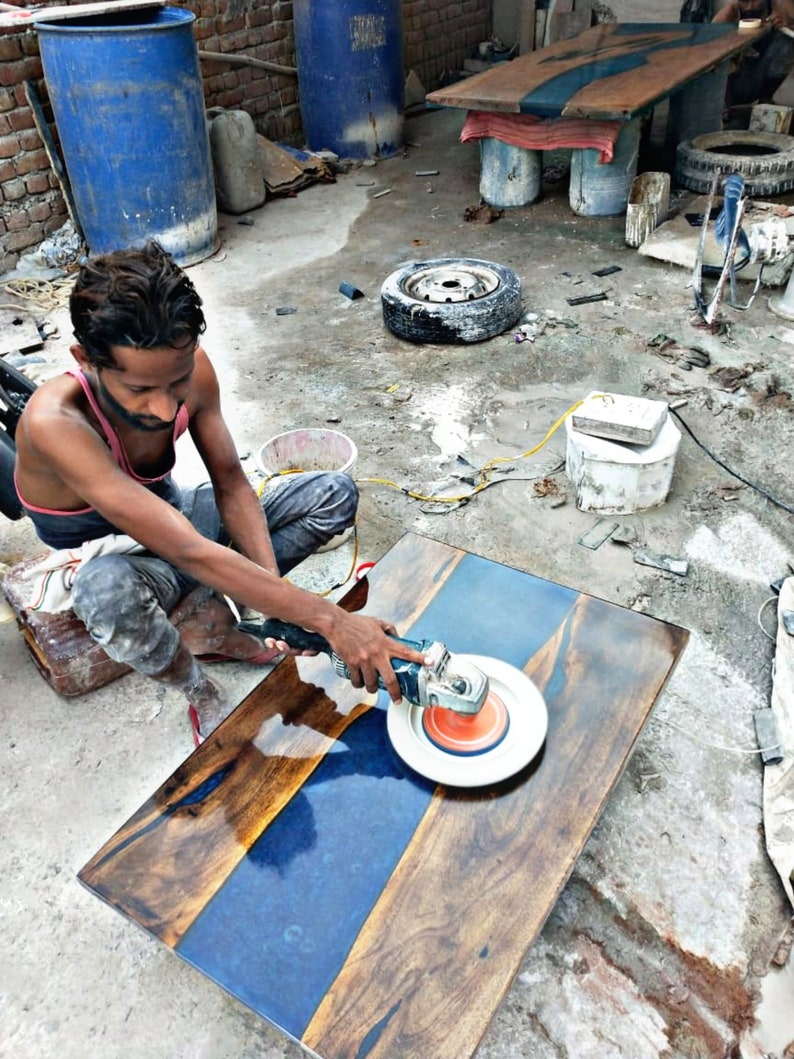 May include: A person uses a power sander to polish a wooden table top with a blue epoxy resin inlay. The table top is made of dark wood with a natural grain pattern.