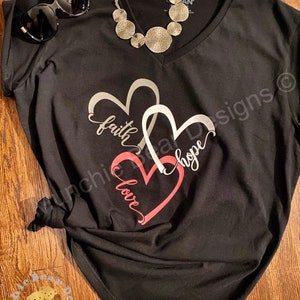 May include: Black v-neck t-shirt with a silver design of two overlapping hearts. The hearts have the words "faith", "hope", and "love" written inside them.