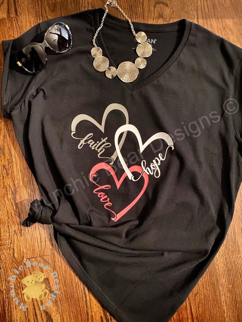 May include: Black v-neck t-shirt with a silver design of two overlapping hearts. The hearts have the words "faith", "hope", and "love" written inside them.