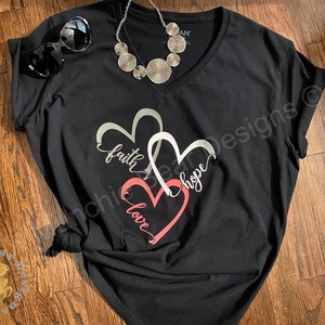 May include: Black v-neck t-shirt with a design of two intertwined hearts. The hearts are silver with the words "faith" and "hope" written inside. The bottom heart is pink with the word "love" written inside.