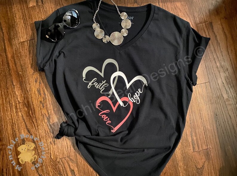 May include: Black v-neck t-shirt with a design of two intertwined hearts. The hearts are silver with the words "faith" and "hope" written inside. The bottom heart is pink with the word "love" written inside.