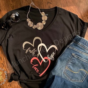 May include: Black v-neck t-shirt with a graphic of three interlocking hearts. The hearts are silver, white, and pink. The words "faith", "hope", and "love" are written inside the hearts. The shirt is paired with blue jeans.