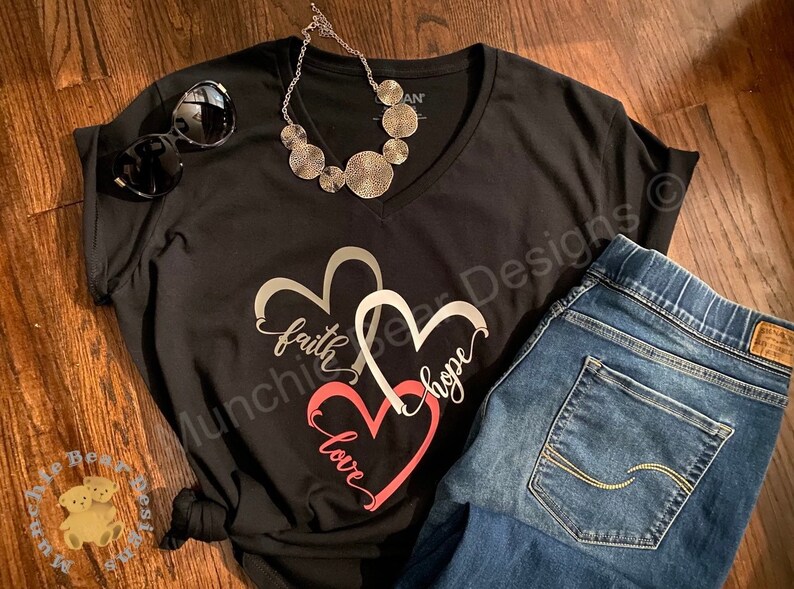 May include: Black v-neck t-shirt with a graphic of three interlocking hearts. The hearts are silver, white, and pink. The words "faith", "hope", and "love" are written inside the hearts. The shirt is paired with blue jeans.