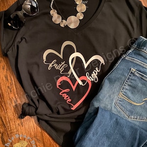 May include: Black v-neck t-shirt with a graphic of three interlocking hearts. The hearts are silver, white, and pink. The words "faith", "hope", and "love" are written inside the hearts. The shirt is paired with blue jeans and a silver necklace.