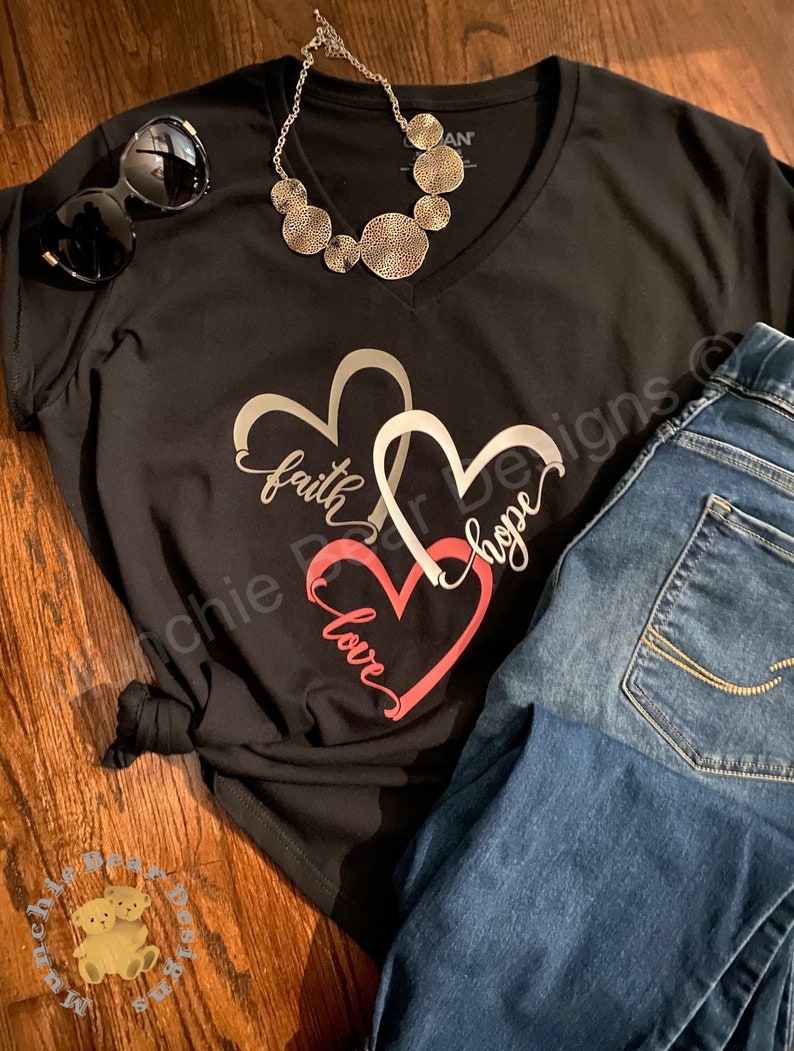 May include: Black v-neck t-shirt with a graphic of three interlocking hearts. The hearts are silver, white, and pink. The words "faith", "hope", and "love" are written inside the hearts. The shirt is paired with blue jeans and a silver necklace.