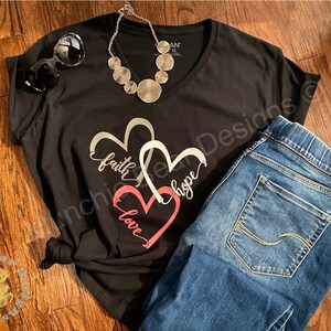 May include: Black t-shirt with a silver and pink heart design. The design features the words "faith", "hope", and "love" in a cursive font. The shirt is paired with blue jeans.