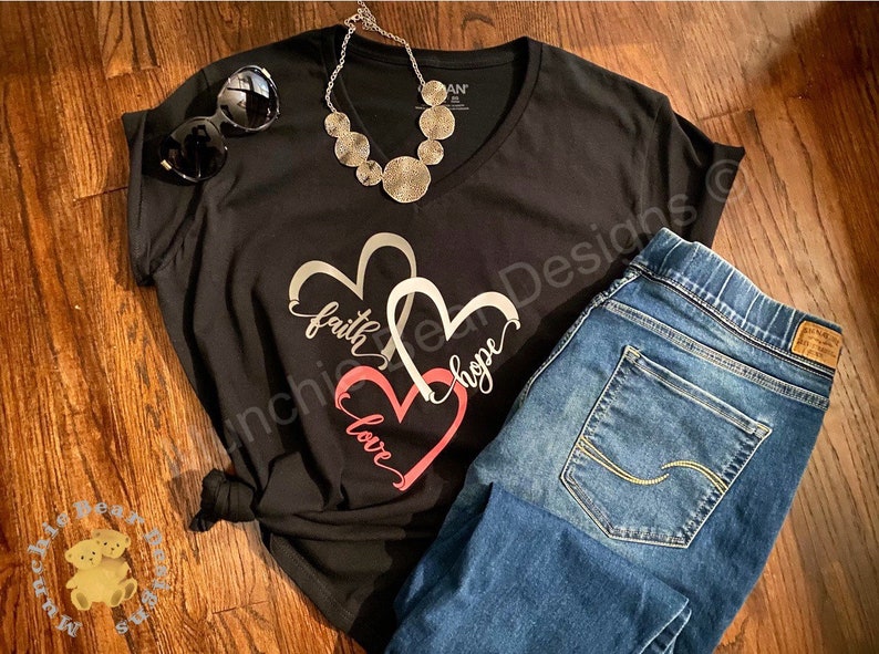 May include: Black t-shirt with a silver and pink heart design. The design features the words "faith", "hope", and "love" in a cursive font. The shirt is paired with blue jeans.
