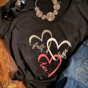 May include: Black v-neck t-shirt with a graphic of three interlocking hearts. The hearts are outlined in silver and filled with pink, gray, and white. The words "faith", "hope", and "love" are written inside the hearts.